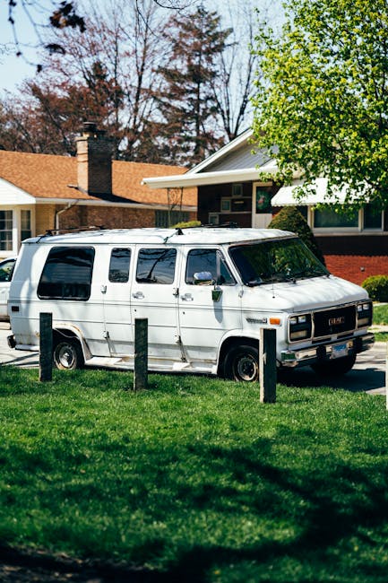 A white vintage GMC van parked on a residential street with a grassy verge in front, surrounded by wooden bollards. The van's sliding door is open, revealing a partially loaded interior used for home relocation, with cardboard boxes, wrapped furniture, and packing materials visible inside. The van is positioned near a suburban house with a pitched roof, large windows, and a chimney, set against a backdrop of trees with green and autumnal foliage. The scene, captured during daylight with clear weather, depicts preparations for furniture transport and moving logistics by a professional removals service, consistent with house moving activities in Grove Park. The image highlights the loading process involving careful placement of household items into the vehicle, supported by the presence of protective equipment such as blankets and straps often used by [COMPANY_NAME].