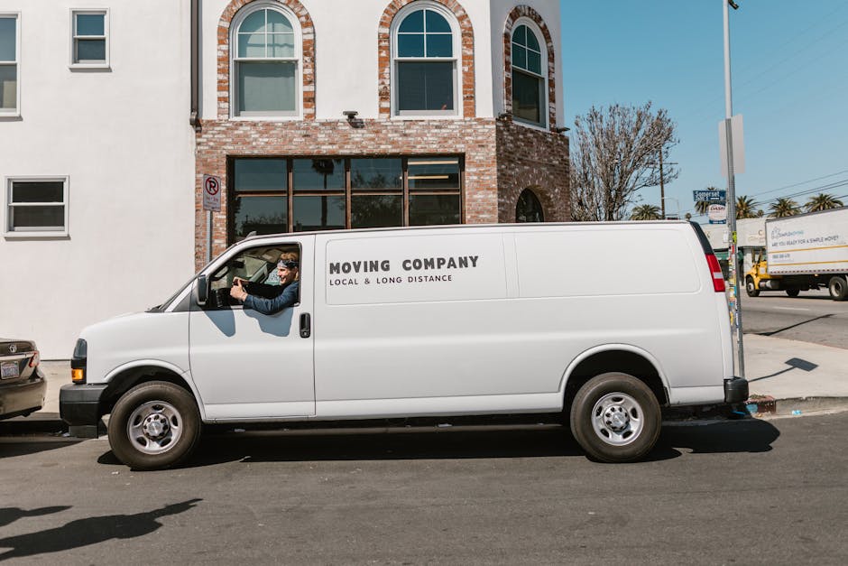A white vintage GMC van parked on a residential street with a grassy verge in front, surrounded by wooden bollards. The van's sliding door is open, revealing a partially loaded interior used for home relocation, with cardboard boxes, wrapped furniture, and packing materials visible inside. The van is positioned near a suburban house with a pitched roof, large windows, and a chimney, set against a backdrop of trees with green and autumnal foliage. The scene, captured during daylight with clear weather, depicts preparations for furniture transport and moving logistics by a professional removals service, consistent with house moving activities in Grove Park. The image highlights the loading process involving careful placement of household items into the vehicle, supported by the presence of protective equipment such as blankets and straps often used by [COMPANY_NAME].