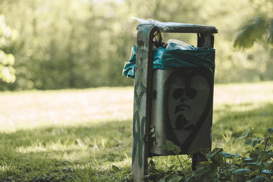 A metal waste bin with spray-painted graffiti depicting a stylised cartoon face, including dark sunglasses, a closed mouth, and a tongue sticking out, is positioned on a grassy area near a property. The bin is partially covered with blue plastic bags and other waste materials, which appear to contain household items or rubbish. Surrounding the bin are green leafy plants, and in the background, there are blurred trees and sunlight filtering through the foliage, indicating an outdoor setting. The scene suggests an area where bulky waste or garden refuse may be temporarily placed, supporting home relocation or clearance activities, as seen in professional removals and furniture transport services, with the bin likely being part of a waste disposal process related to house moving or renovation in Grove Park. Man with Van Grove Park offers removals and waste clearance options connected to house moves, ensuring proper handling of bulk waste.