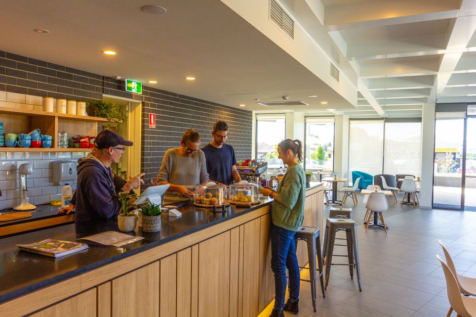 The image shows the interior of a café with a spacious layout featuring multiple wooden tables and chairs, some with patterned cushions, arranged neatly across a tiled floor. The walls are painted in