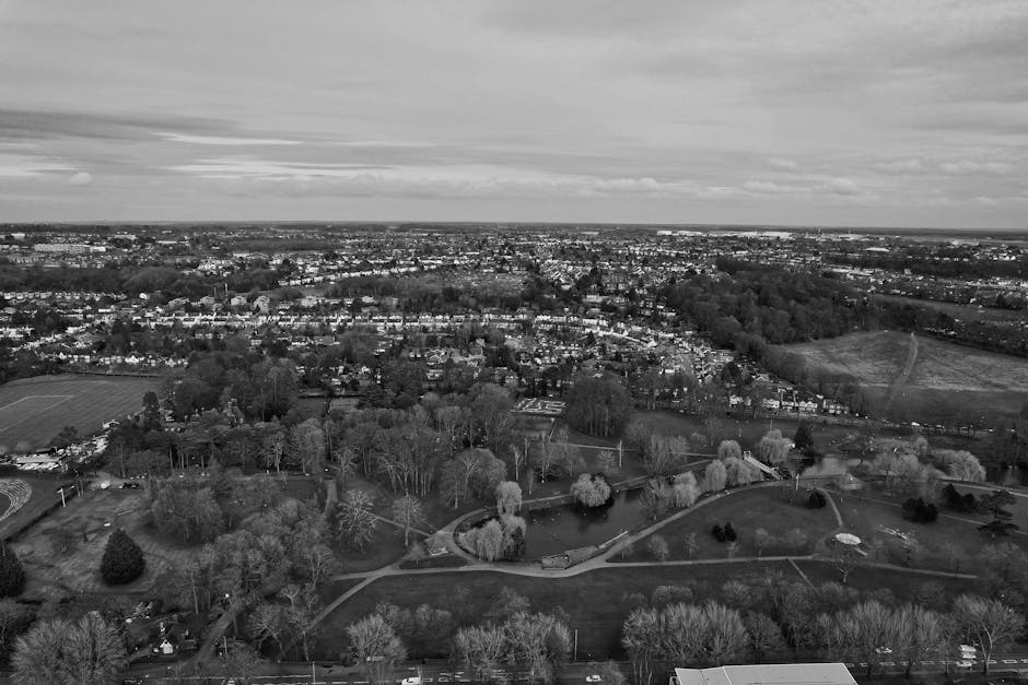 Aerial black and white photograph of a suburban residential area showing a park with winding pathways, numerous trees, and small bodies of water in the foreground. The park is surrounded by low-rise houses, with some properties featuring visible gardens. Beyond the park, the landscape extends into a densely populated area with numerous houses, roads, and scattered greenery, all under a cloudy sky. In the context of home relocation, this image illustrates the outdoor environment where furniture or boxes may be loaded into a van for moving services. The scene, captured during daytime with diffuse lighting, emphasizes the residential setting and infrastructure involved in the furniture transport and packing and moving process. Man with Van Grove Park might utilize such an area during stair and lift solutions for house removals, supporting logistical planning around outdoor access points and transportation routes.