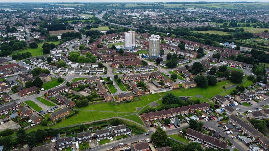 An aerial view of a residential estate showing multiple brick-built apartment buildings with dark tiled roofs, surrounded by green lawns and trees, with a network of paved roads and parking areas filled with cars. The image captures the scene during daylight with natural lighting, highlighting the well-maintained landscaping and spacious outdoor areas. This setting reflects a typical environment where home relocation and furniture transport services by Man with Van Grove Park could be involved, illustrating potential logistics such as the loading and unloading of packing materials, boxes, and furniture from vans parked nearby within the estate premises, as part of stair and lift solution arrangements for GROVE PARK's Chinbrook Meadows estate moves.