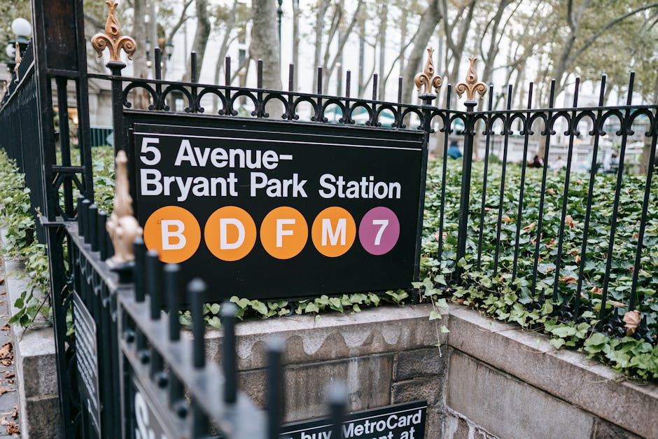 A close-up view of a metal gate surrounding the entrance to Grove Park station, with a black and white sign displaying station details including the address '5 Avenue-Bryant Park Station' and the London Underground symbols for lines B, D, F, M, and 7 in orange and purple circles. The sign is mounted on a stone and concrete base with green ivy growing along the edges, and behind the gate, there are trees and a glimpse of parked vehicles. The scene suggests an outdoor setting during daytime, relevant to house removals and moving logistics, emphasizing the importance of station accessibility and familiar landmarks during home relocation or furniture transport.