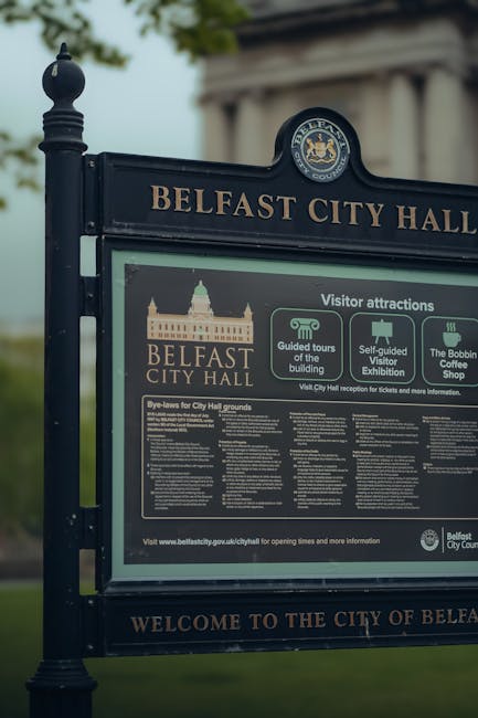A large black metal signboard displaying information about Belfast City Hall, located outdoors against a background of green trees and cloudy sky. The sign features the Belfast City Hall crest at the top, with the inscription 'Belfast City Hall' below it, followed by icons and details about visitor attractions such as guided tours, self-guided audio, and the Bobbin Coffee Shop. The lower section contains extensive text on bye-laws for City Hall grounds. The sign is positioned on a paved area and is part of a house removal or relocation context, similar to services provided by Man with Van Grove Park. The overall scene emphasizes the informational and organizational aspects of city landmarks relevant to residential or commercial moves involving Belfast City Hall.