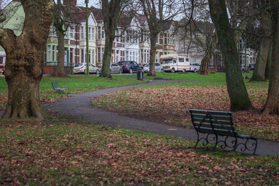 A quiet residential park area with a winding paved walkway surrounded by large trees with textured bark and green, orange, and brown fallen leaves covering the grass. Two empty metal park benches with decorative curved armrests and slatted seats and backs are positioned along the pathway—one prominently in the foreground on the right and the other further back on the left. In the background, there is a row of Victorian-style terraced houses with bay windows, painted in pastel colors, and multiple parked cars including a white van and a few smaller vehicles, indicating street parking adjacent to the park. The scene is illuminated by natural daylight, with overcast sky conditions, and the environment suggests a peaceful setting suitable for a home relocation or house move, supported by the proximity of residential properties and parking arrangements. The presence of the park benches and the pathway hints at a community area often used during the logistics of packing and moving furniture to or from nearby residences, with Man with Van Grove Park providing related removals services.