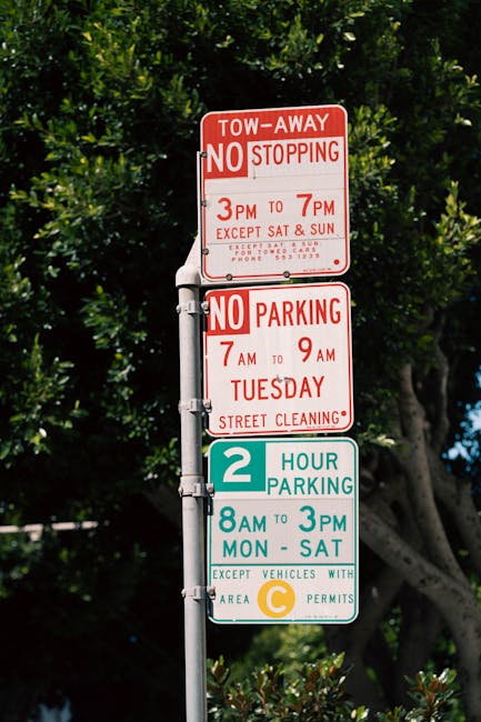 A vertical metal signpost displaying multiple parking and stopping regulations, including a red and white 'Tow-Away No Stopping' notice from 3 PM to 7 PM, with additional restrictions for no parking from 7 AM to 9 AM on Tuesdays for street cleaning. Below these, a green and white sign indicates a two-hour parking limit from 8 AM to 3 PM, Monday to Saturday, for vehicles with a C area permit. The signpost is set against a backdrop of dense green trees with sunlight filtering through the leaves, situated along a paved pavement. This signage is relevant for residents and visitors planning to execute home relocations or moving activities within Grove Park, highlighting restrictions to avoid parking fines during furniture transport or loading and unloading processes, as managed by Man with Van Grove Park.