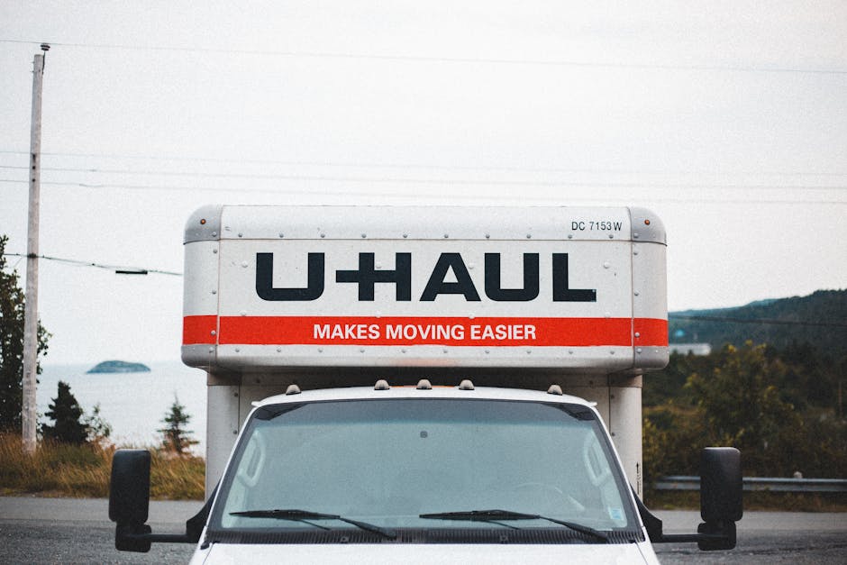 Close-up view of the front of a white U-Haul moving van parked outdoors under an overcast sky, with the large cargo area visible above the cab. The cargo area displays the black U-Haul logo and a red stripe with the slogan 'MAKES MOVING EASIER.' Surrounding the vehicle are blurred trees, power lines, and distant hills, suggesting a suburban or rural area. Inside the scene, the van's windshield and side mirrors are visible, with the front part of a vehicle reflecting on the glass. The parking space appears on a paved surface, and the setting indicates preparations for house relocation or furniture transport, consistent with professional removal and moving services provided by companies like Man with Van Grove Park. The overall lighting is natural, with diffuse daylight typical of an overcast day, emphasizing the functional and logistical context of loading or transporting household items.