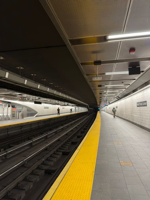 The image shows an underground train station platform with a wide, empty tiled surface and a yellow tactile paving strip along the edge for safety. The platform is illuminated by overhead fluorescent 